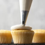 A piping tip pipes vanilla buttercream frosting over a vanilla cupcake, next to more cupcakes on a wire rack.