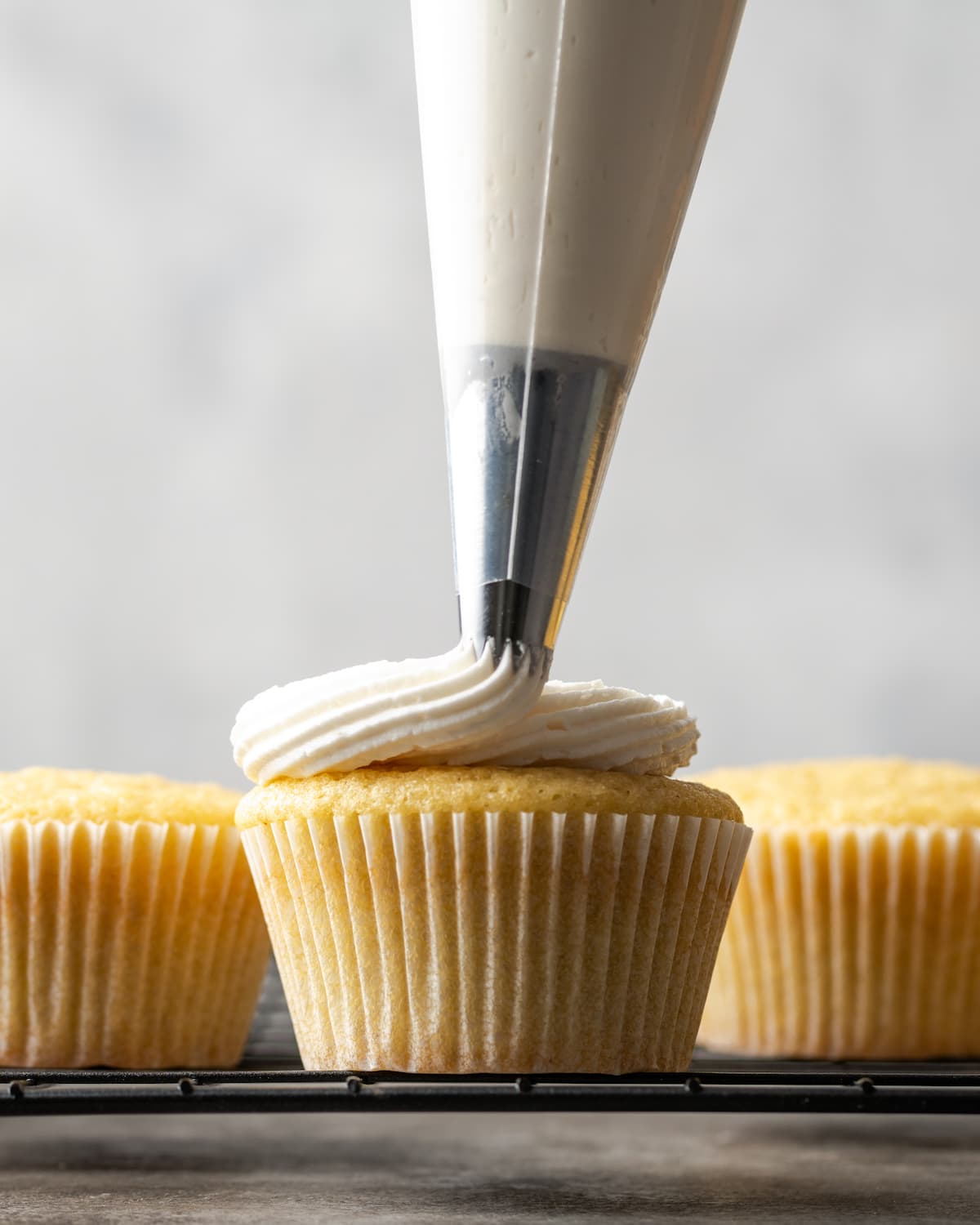 A piping tip pipes vanilla buttercream frosting over a vanilla cupcake, next to more cupcakes on a wire rack.