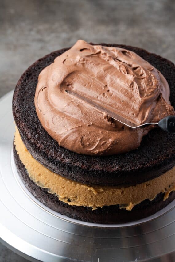 An offset spatula being used to spread chocolate frosting generously over top of a peanut butter and chocolate layer cake.