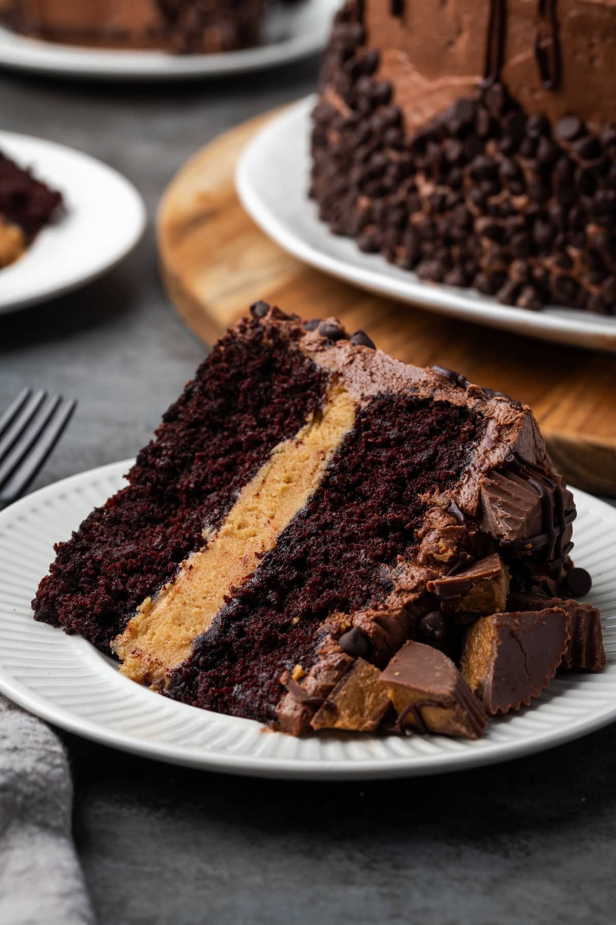 This Reese's cake is a moist chocolate cake filled with an insane amount of peanut butter frosting, topped with heaps of peanut butter cups! A slice of Reese's cake on a white plate with the rest of the cake in the background.
