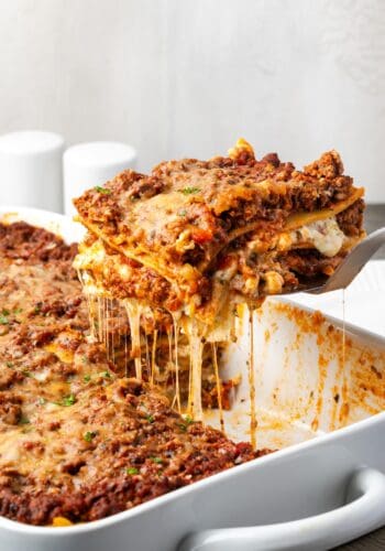 A slice of the best lasagna being lifted from the rest of the lasagna in a ceramic baking dish.