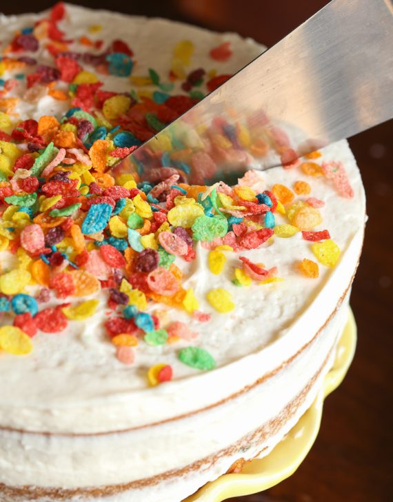 Overhead view of a knife cutting into a frosted cake topped with fruity pebbles cereal