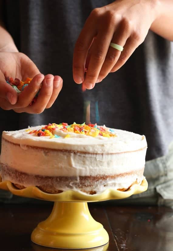 Fruity pebbles cereal being sprinkled on top of a frosted layer cake on a yellow cake stand