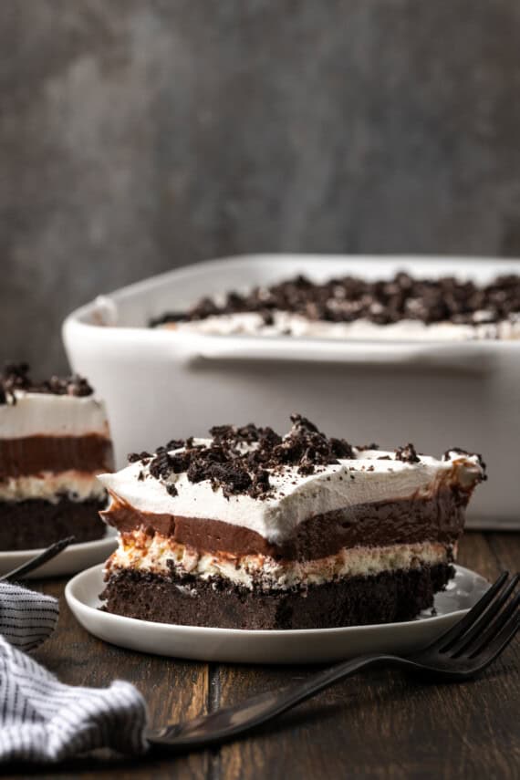 A slice of chocolate lasagna on a plate with the rest of the dessert in a casserole dish in the background.