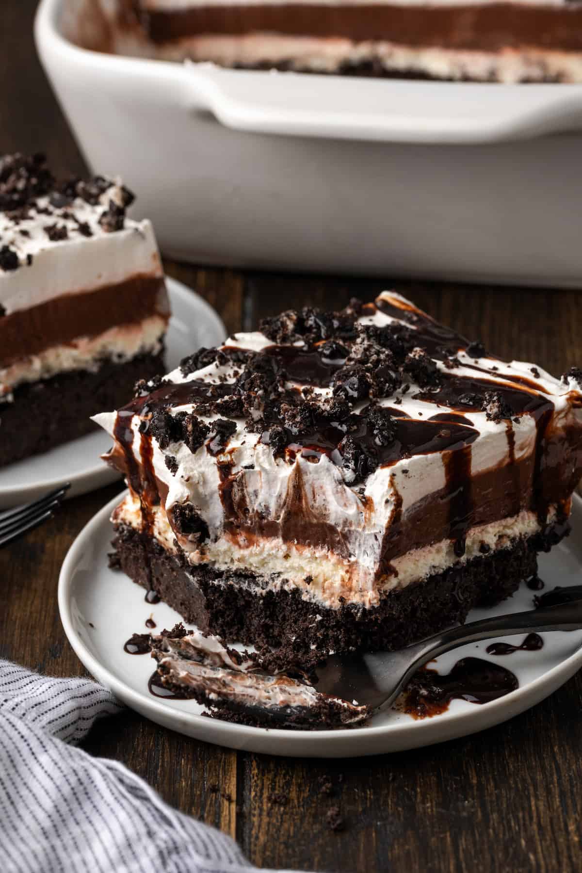 A slice of chocolate lasagna on a plate, topped with chocolate sauce with a forkful missing from the corner.