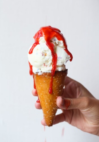 A hand holding a vanilla ice cream cone dripping with strawberry syrup.