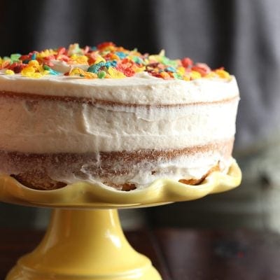 Side view of frosted Cereal Milk Cake topped with Fruity Pebbles on a yellow cake stand