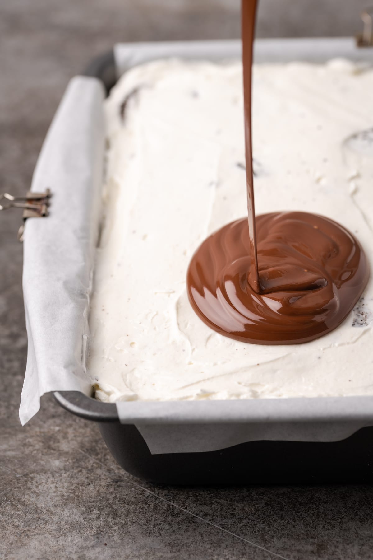 Melted chocolate drizzles and pools on top of the frosting layer of Oreo brownies in a baking pan.