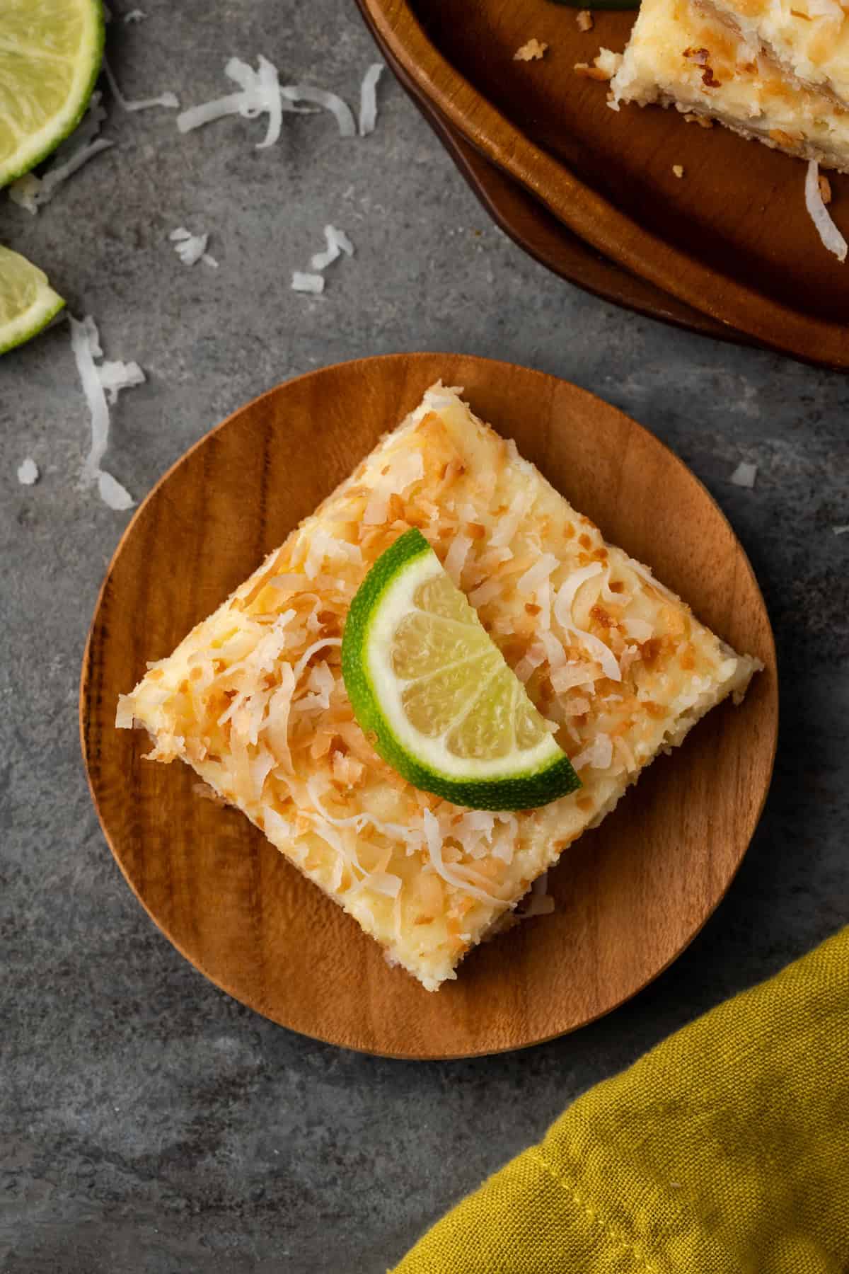 Overhead view of a coconut lime cheesecake bar topped with toasted coconut and a lime wedge on a round wooden plate.