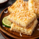 Coconut lime cheesecake bars stacked on a round wooden plate next to a lime wedge.