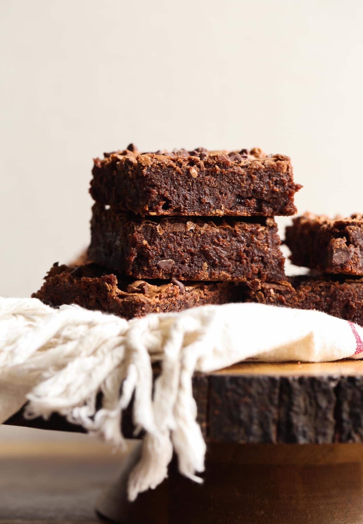 Side view of three fudgy brownies stacked on top of a cloth on a wooden cake stand.