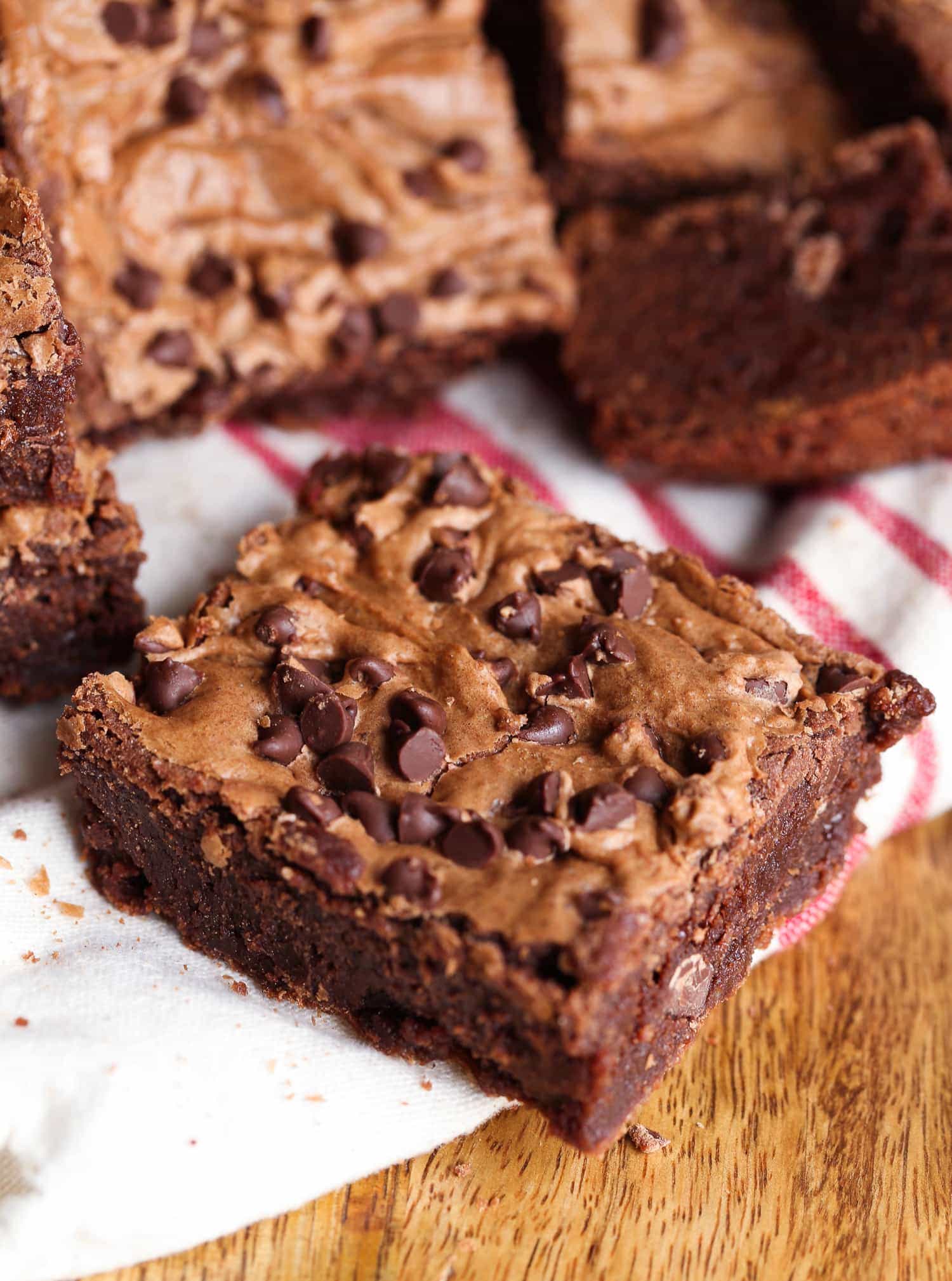 Close up of a fudgy brownie with a crackly top topped with chocolate chips, with more brownies in the background.