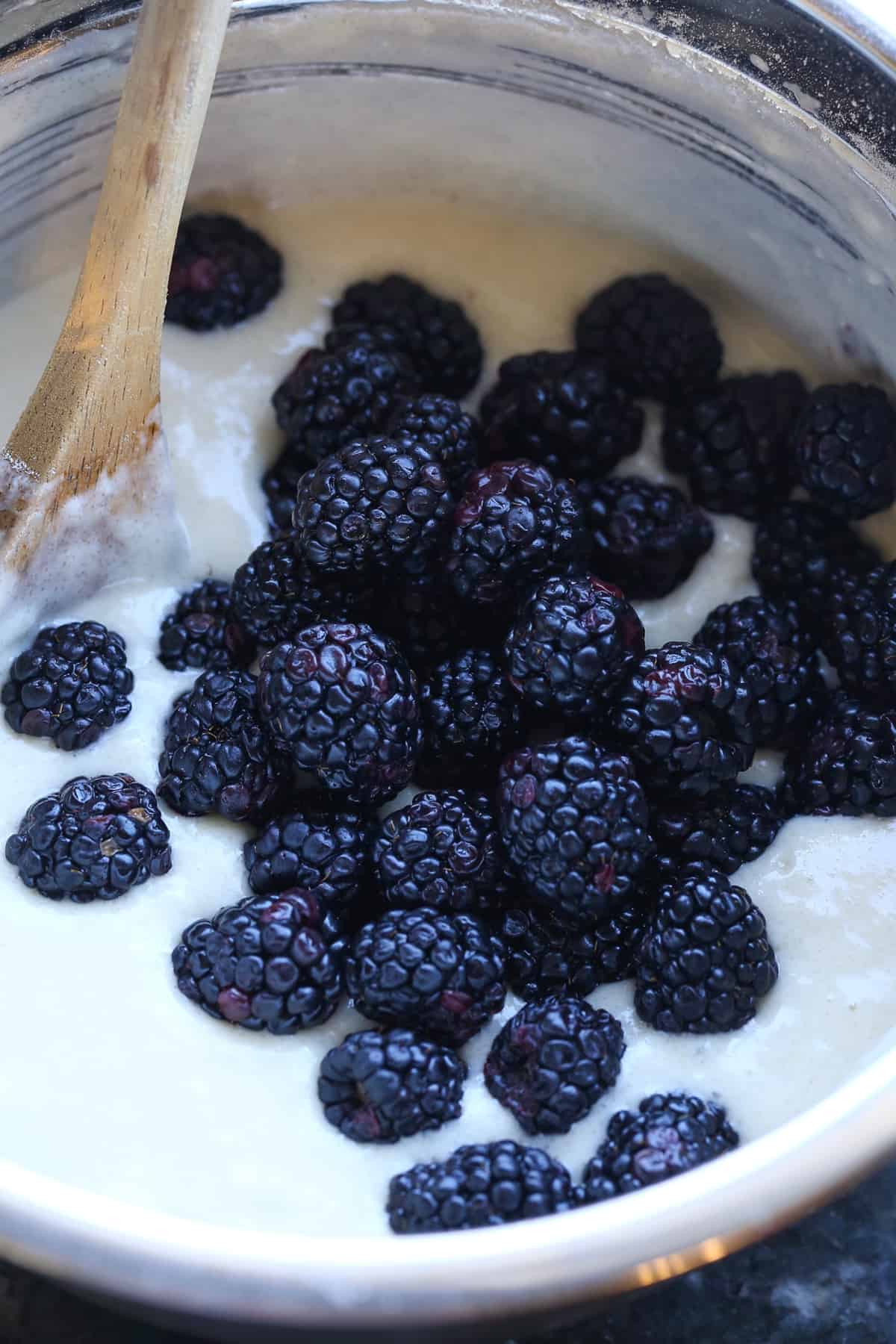 Blackberries being added to cake batter in a bowl with a wooden spoon.
