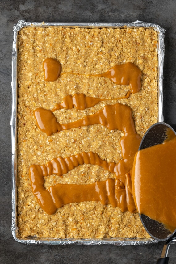 Caramel topping is poured over the dough for caramel oat bars in a sheet pan.