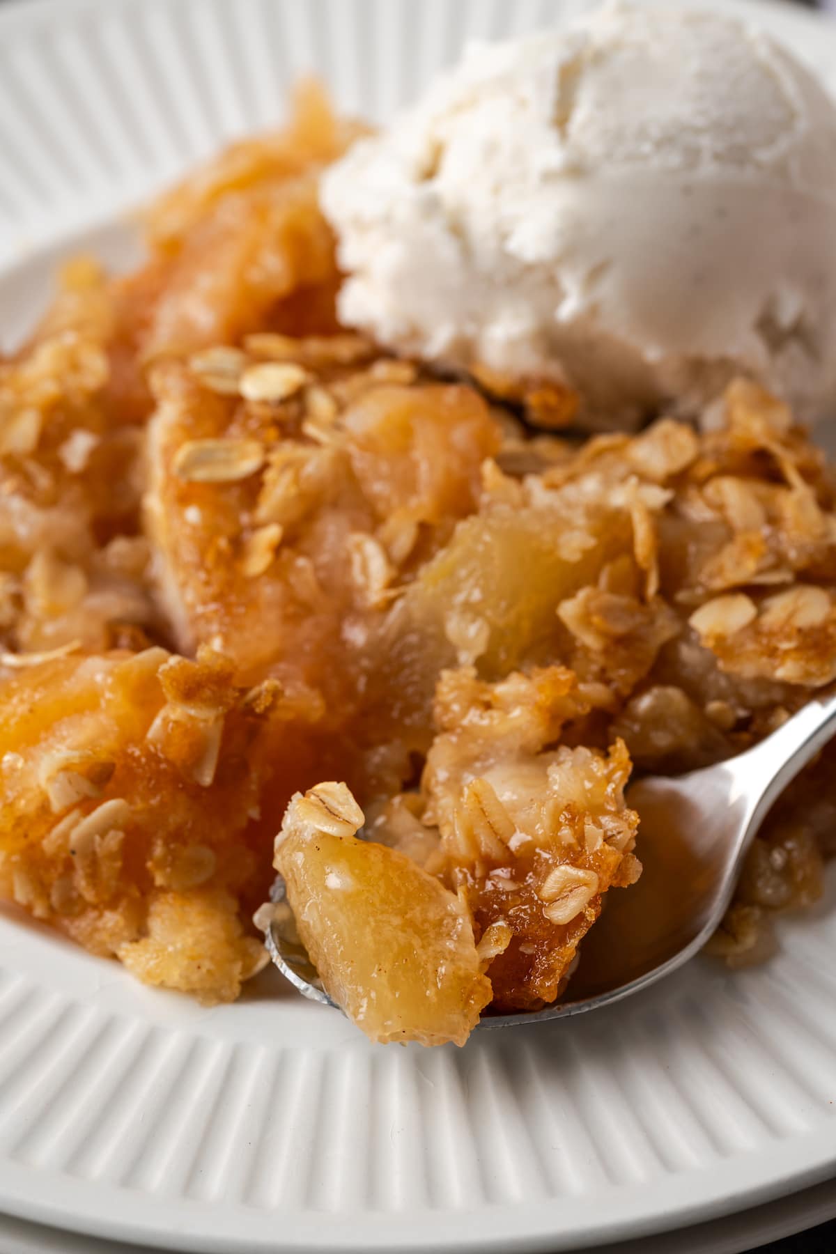 Close up of a serving of apple dump cake on a white plate topped with a scoop of ice cream, next to a spoon.