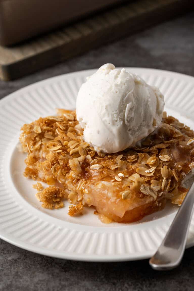 A serving of apple dump cake on a white plate topped with a scoop of ice cream, next to a spoon.