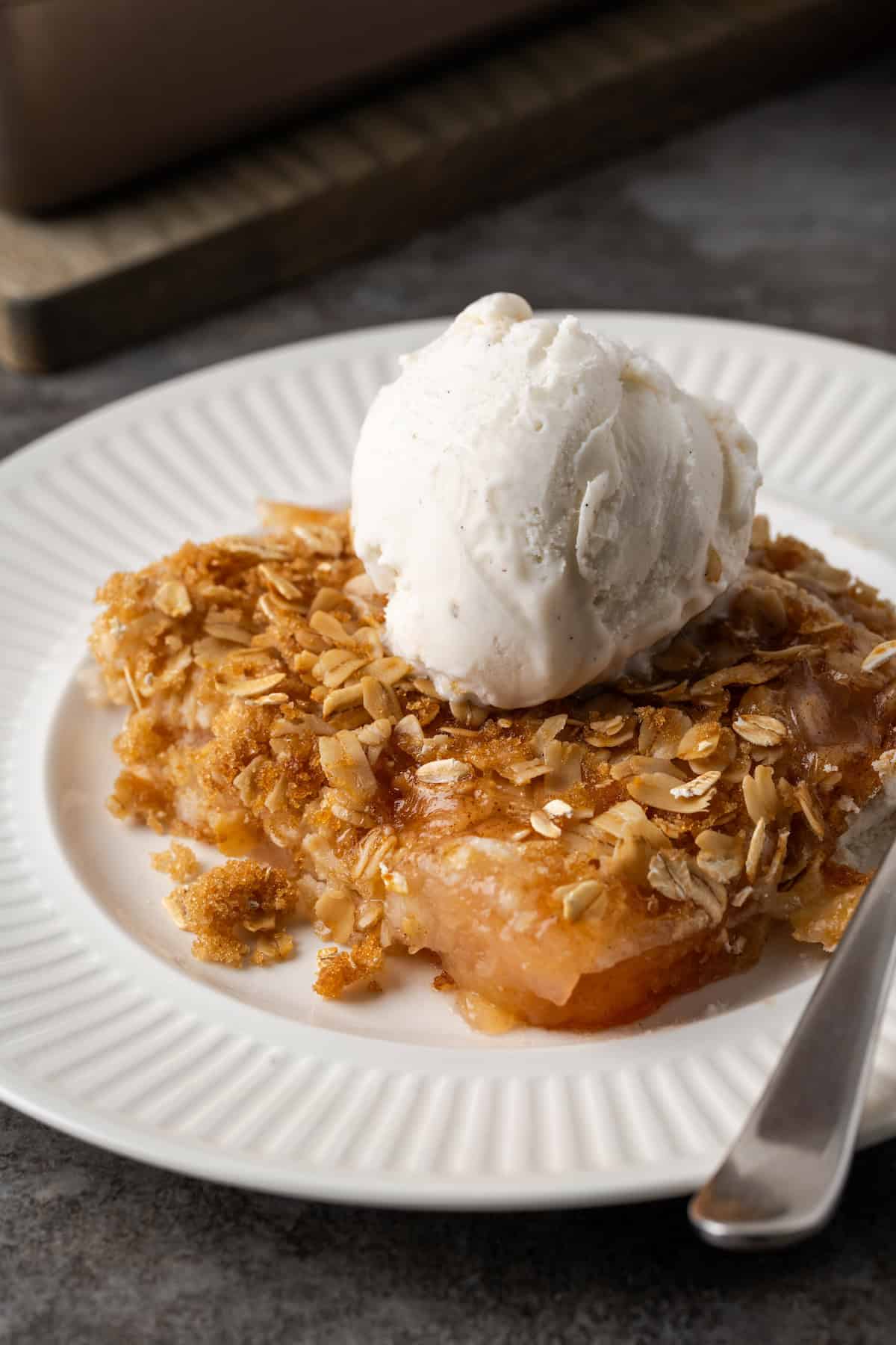 A serving of apple dump cake on a white plate topped with a scoop of ice cream, next to a spoon.