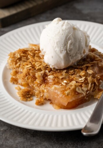 A serving of apple dump cake on a white plate topped with a scoop of ice cream, next to a spoon.