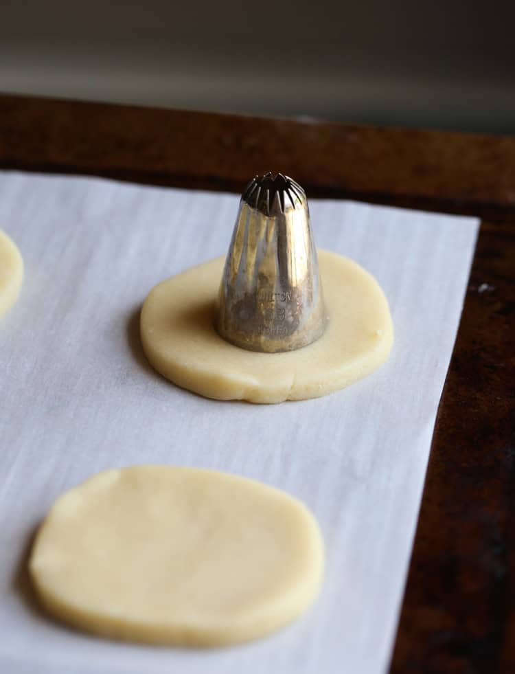 The back of a round piping tip is used to cutout the center of a vanilla cookie dough circle on a parchment lined baking sheet.