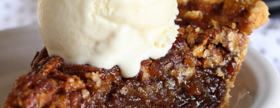 A slice of pecan pie on a white plate topped with a scoop of vanilla ice cream.
