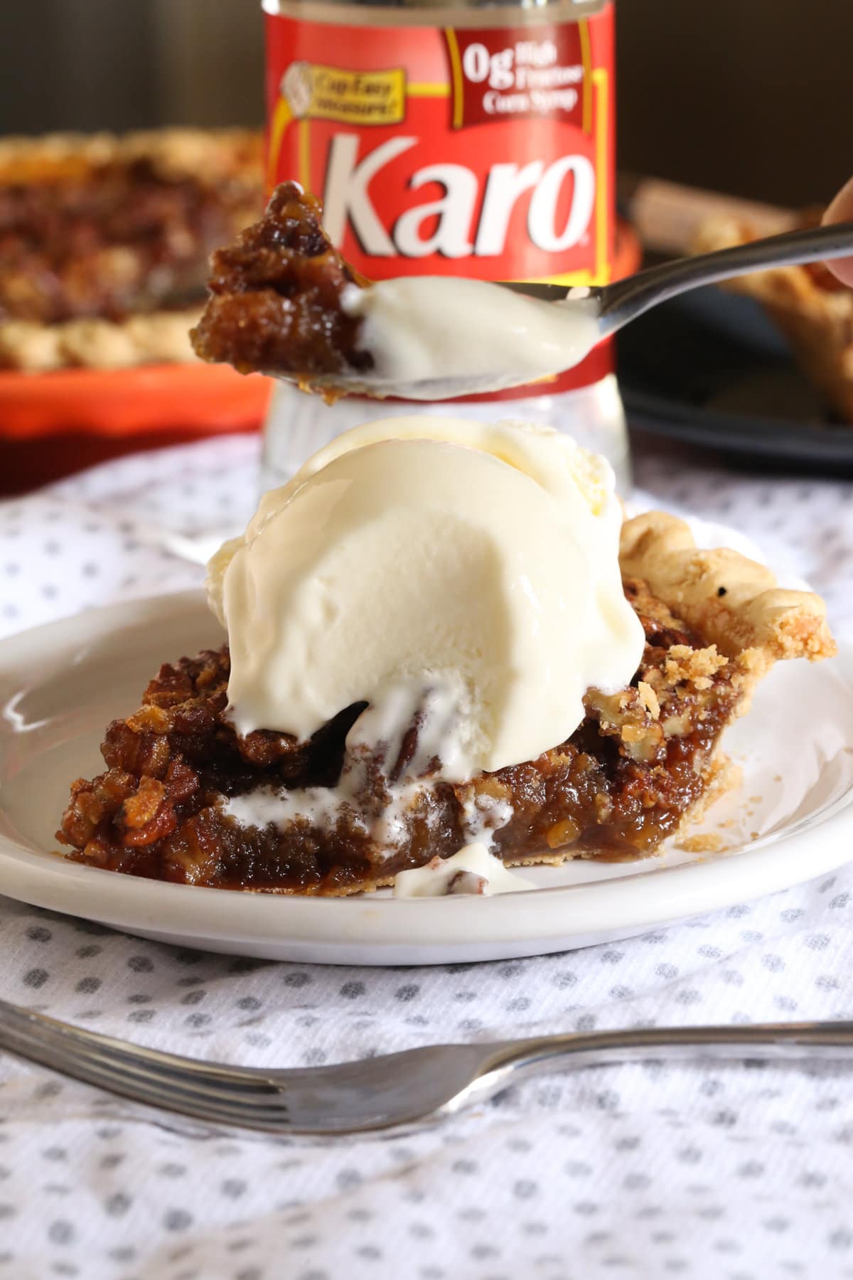 A slice of pecan pie on a plate with a bottle of Karo syrup in the background and a scoop of ice cream on top with a spoon taking a bite