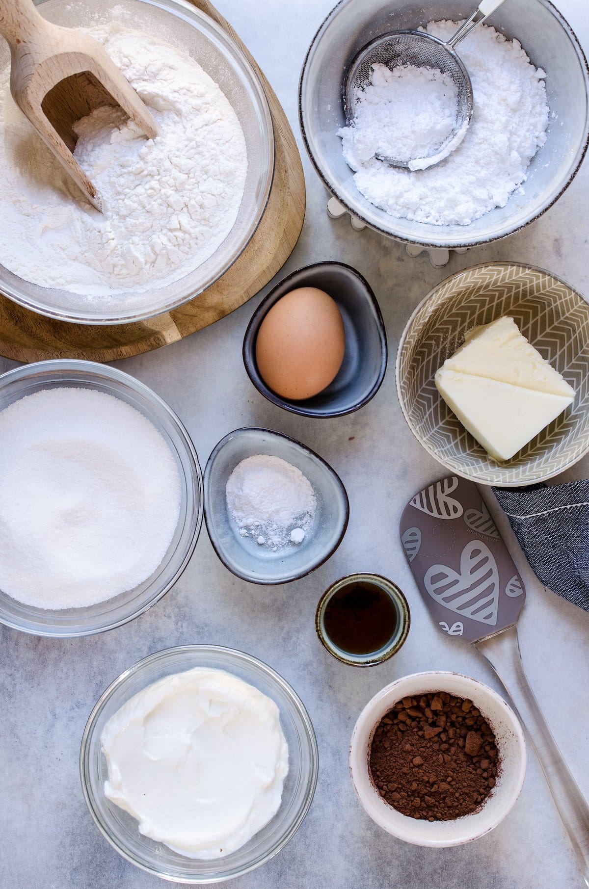The ingredients for black and white cookies: cake flour, powdered sugar, butter, egg, vanilla, sugar, salt, buttermilk, and cocoa powder.