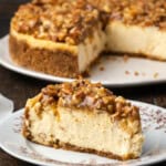 A slices of pecan pie cheesecake on a plate with the rest of the cheesecake in the background.