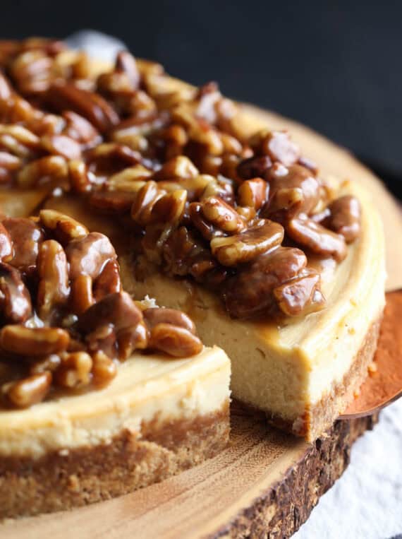 A slice is lifted from a Pecan Pie Cheesecake on a wooden platter.