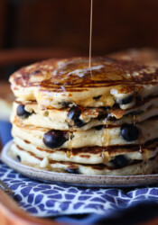 A stack of blueberry pancakes on a plate, being drizzled with maple syrup.