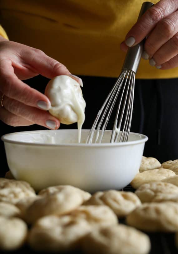 A hand dipping a lemon cream cheese cookie into a bowl of glaze, letting the excess drip off.