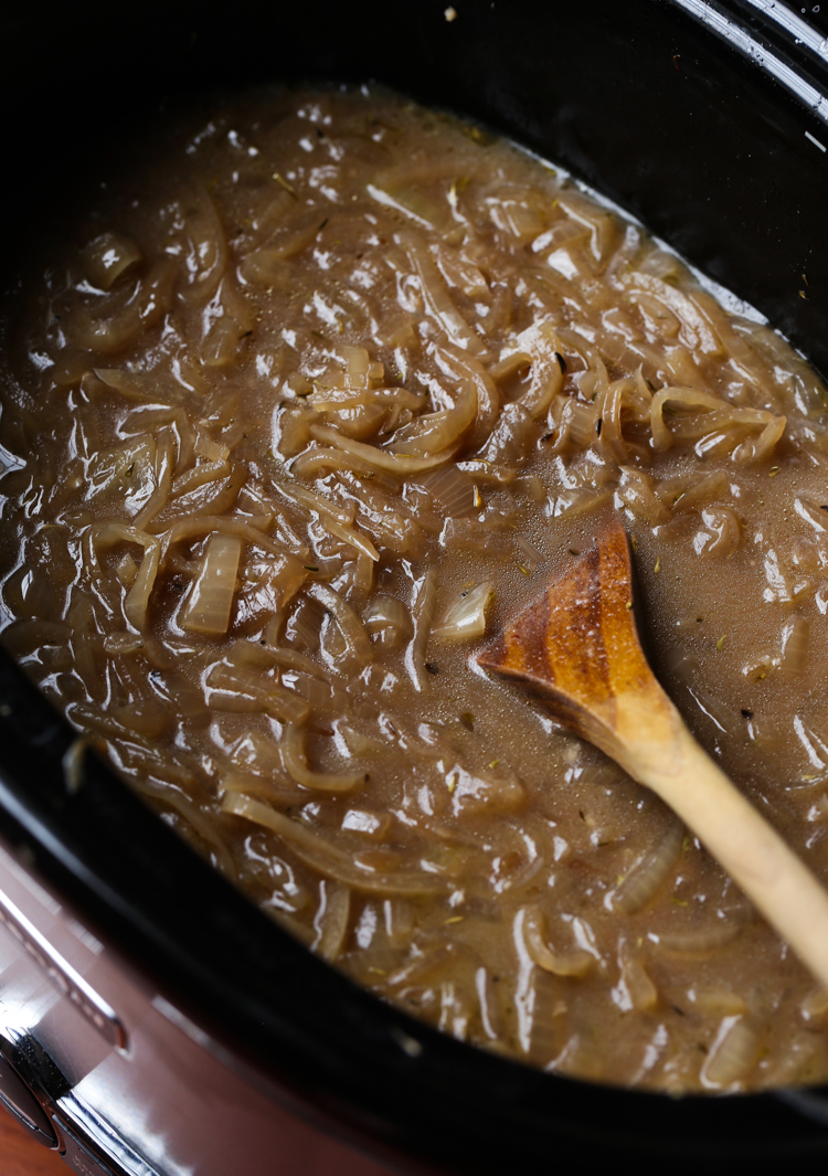 Caramelized onions being stirred together with beef broth, herbs, and wine in the bowl of a slow cooker.