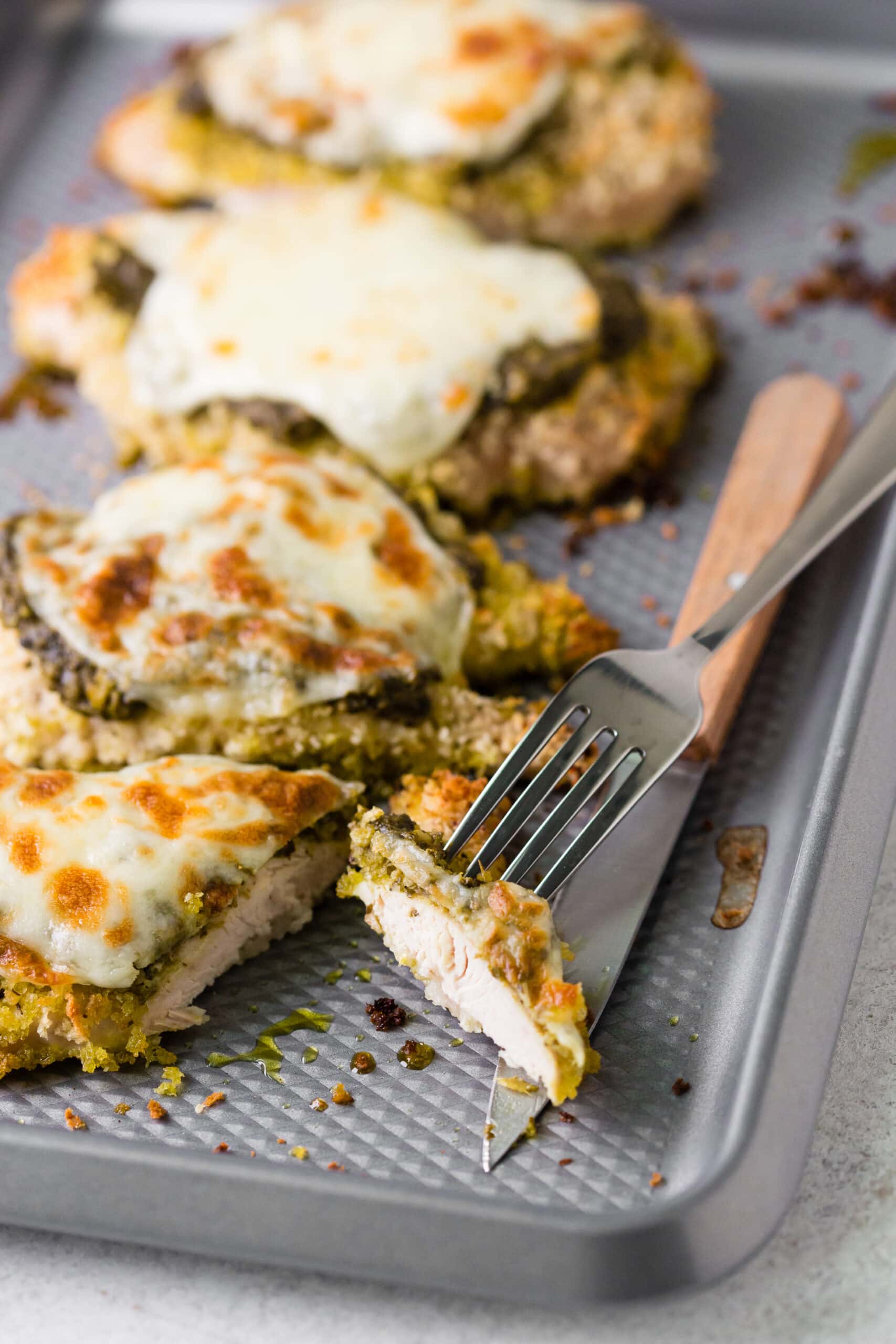 Baked pesto chicken breasts topped with cheese on a baking pan, next to a fork and knife.