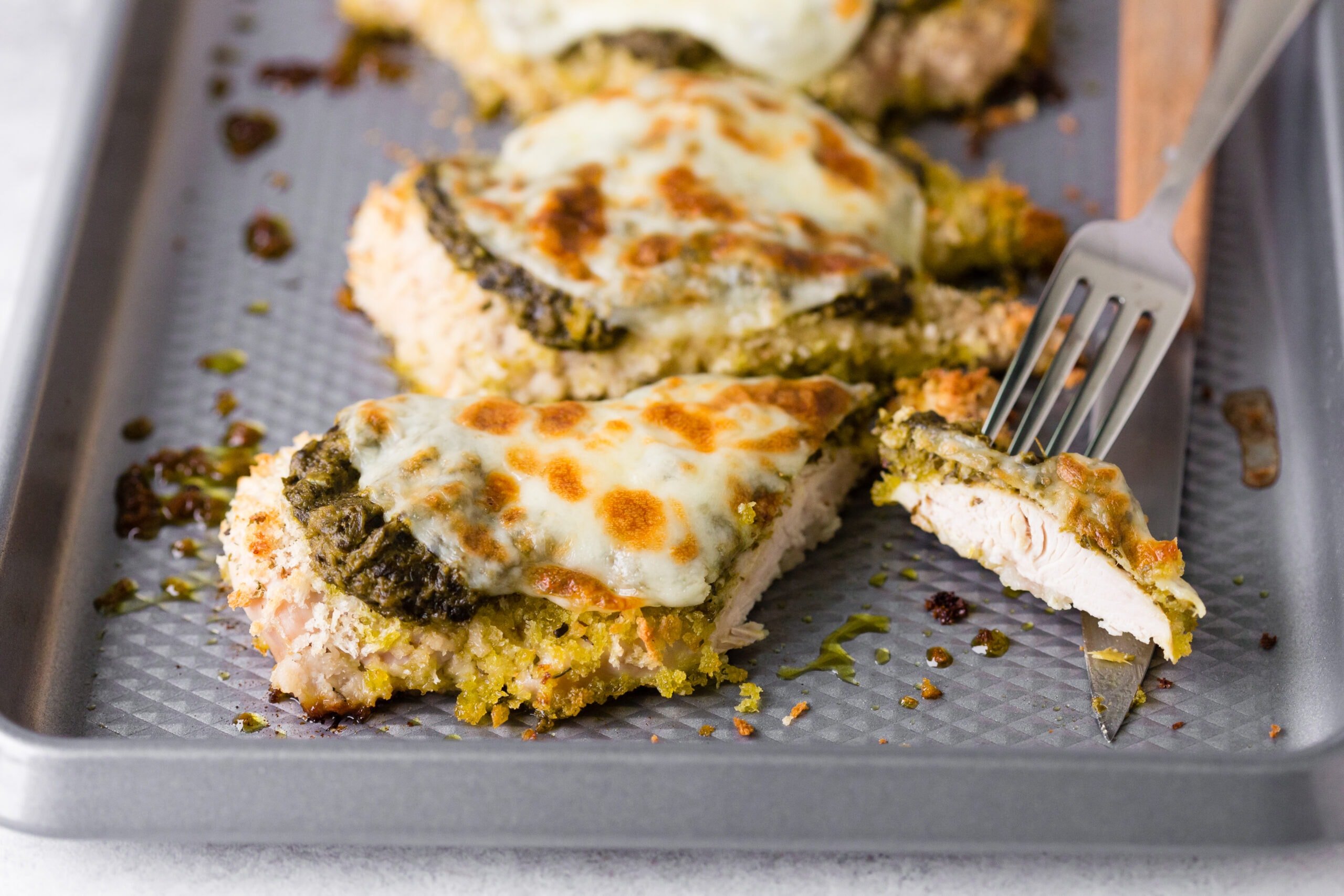 A fork picking up a piece of pesto chicken next to cheesy pesto chicken breasts on a baking pan.