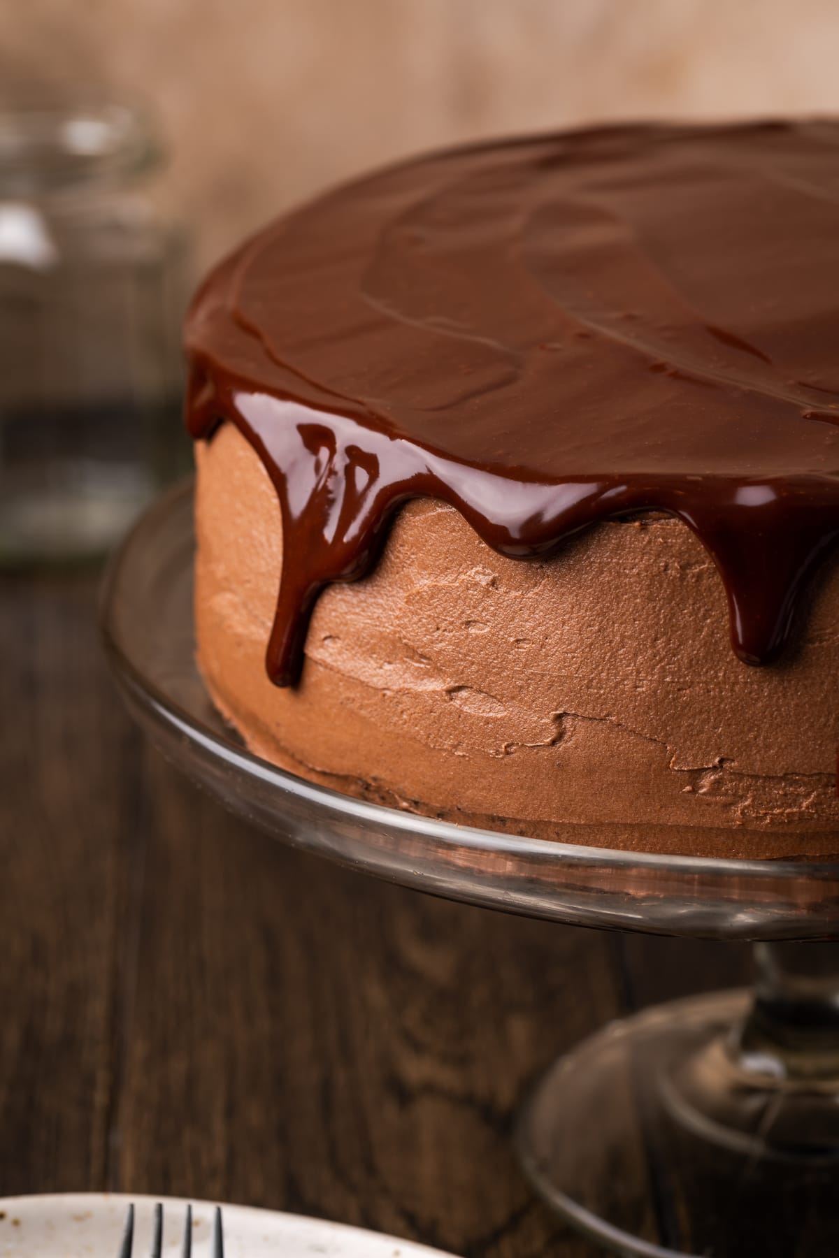 Close up of a frosted chocolate layer cake topped with chocolate ganache on a cake stand.