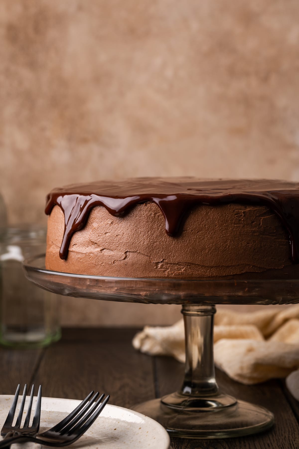 A frosted chocolate layer cake topped with chocolate ganache on a cake stand, next to a white plate with two forks.