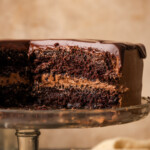 A frosted chocolate layer cake topped with chocolate ganache on a cake stand, with a large slice of cake missing.