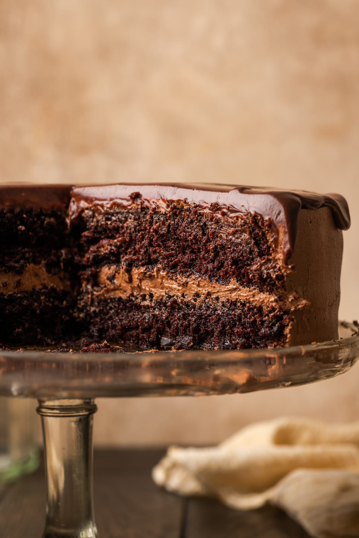 A frosted chocolate layer cake topped with chocolate ganache on a cake stand, with a large slice of cake missing.