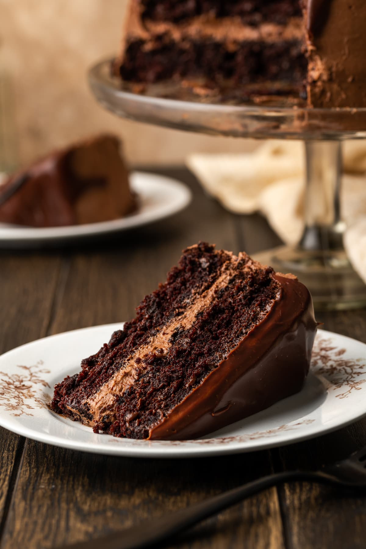 A slice of perfect chocolate layer cake on a white plate next to a fork, with the full cake on a cake stand in the background.