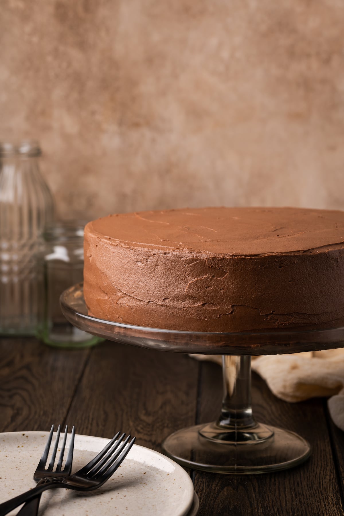 A frosted chocolate layer cake on a cake stand, next to a white plate with two forks.
