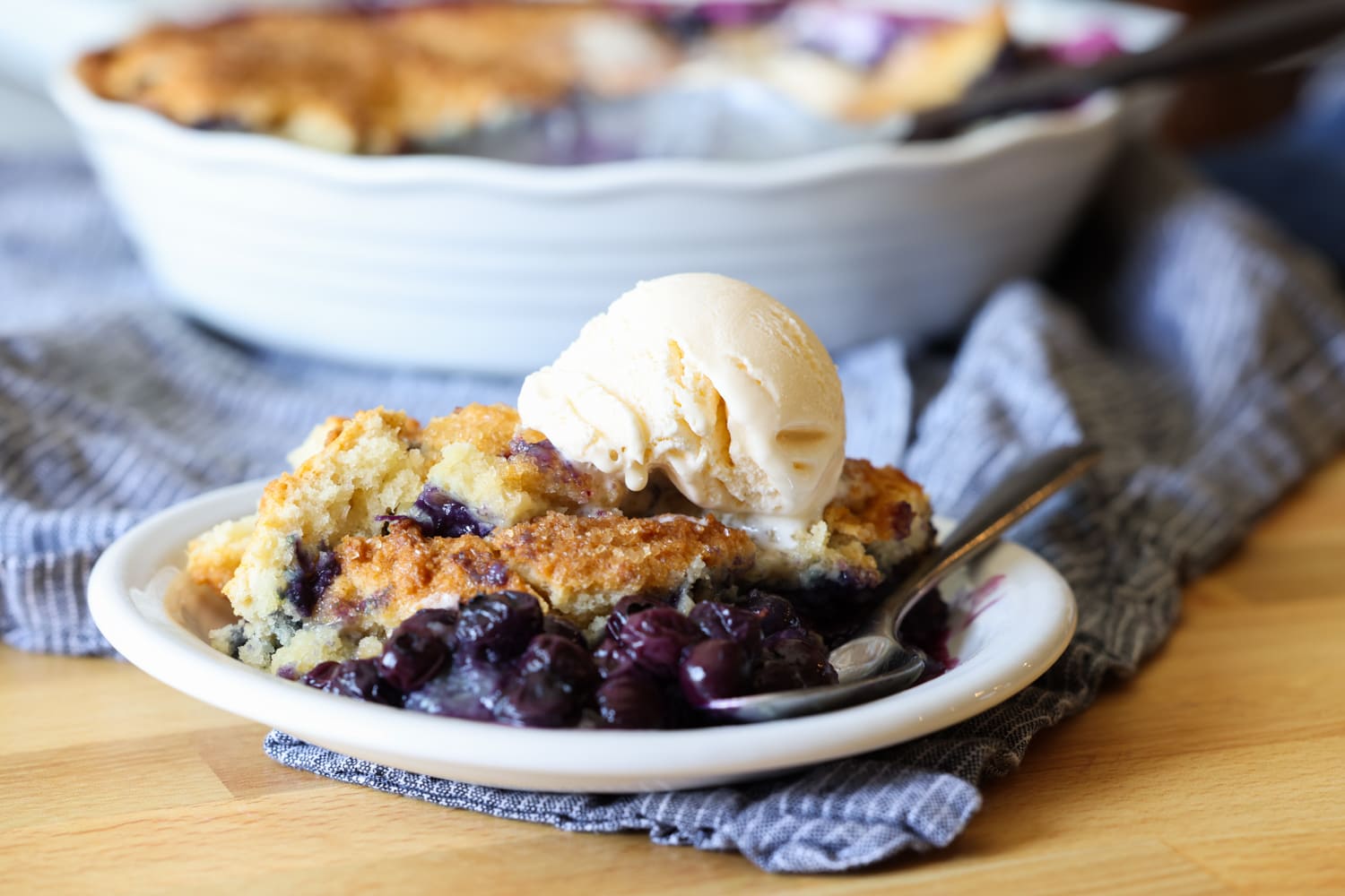 A piece of blueberry cobbler on a white plate with a scoop of ice cream