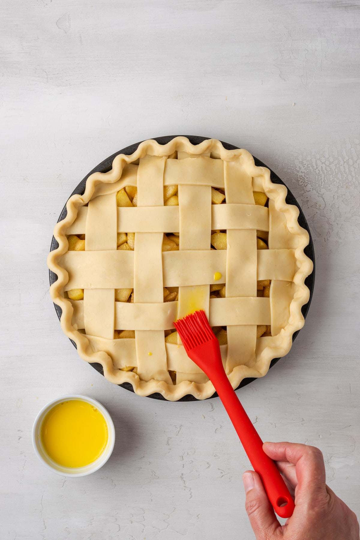 A red rubber brush is used to brush egg wash over an unbaked apple pie with a lattice top next to a small bowl of egg wash.