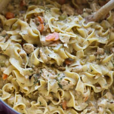 A wooden spoon stirring a large pot of chicken and noodles with carrots and seasoning