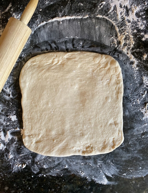 Yeasted dough rolled out into a square on a floured surface, next to a rolling pin.