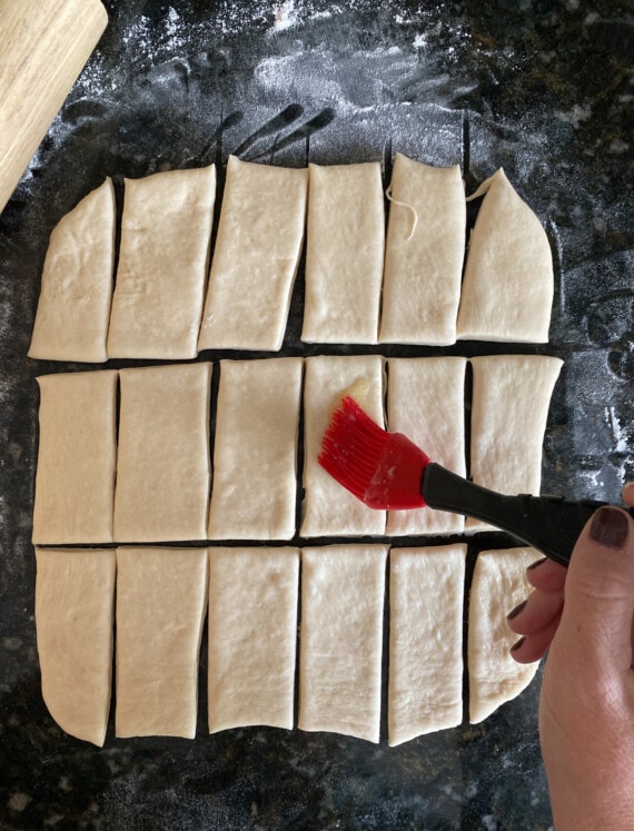 A hand uses a rubber basting brush to brush melted butter over yeasted dough cur into smaller rectangles.