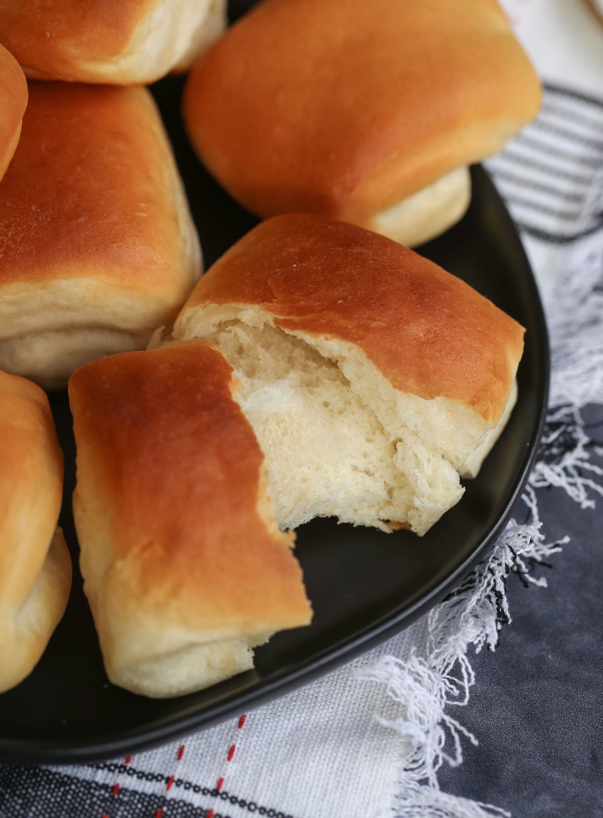 Overhead view of a Parker House roll torn in two on a black plate, next to more rolls.