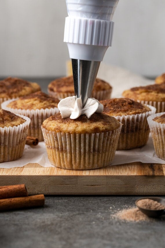 A piping tip pipes cinnamon frosting onto a snickerdoodle cupcake, with more unfrosted cupcakes in the background.