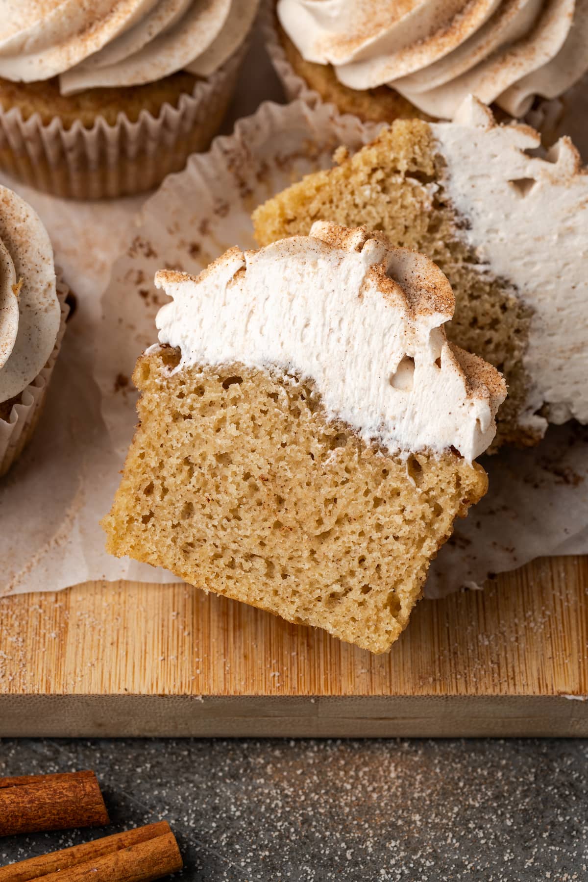 A frosted snickerdoodle cupcake cut in half on a wooden cutting board, next to more frosted cupcakes.