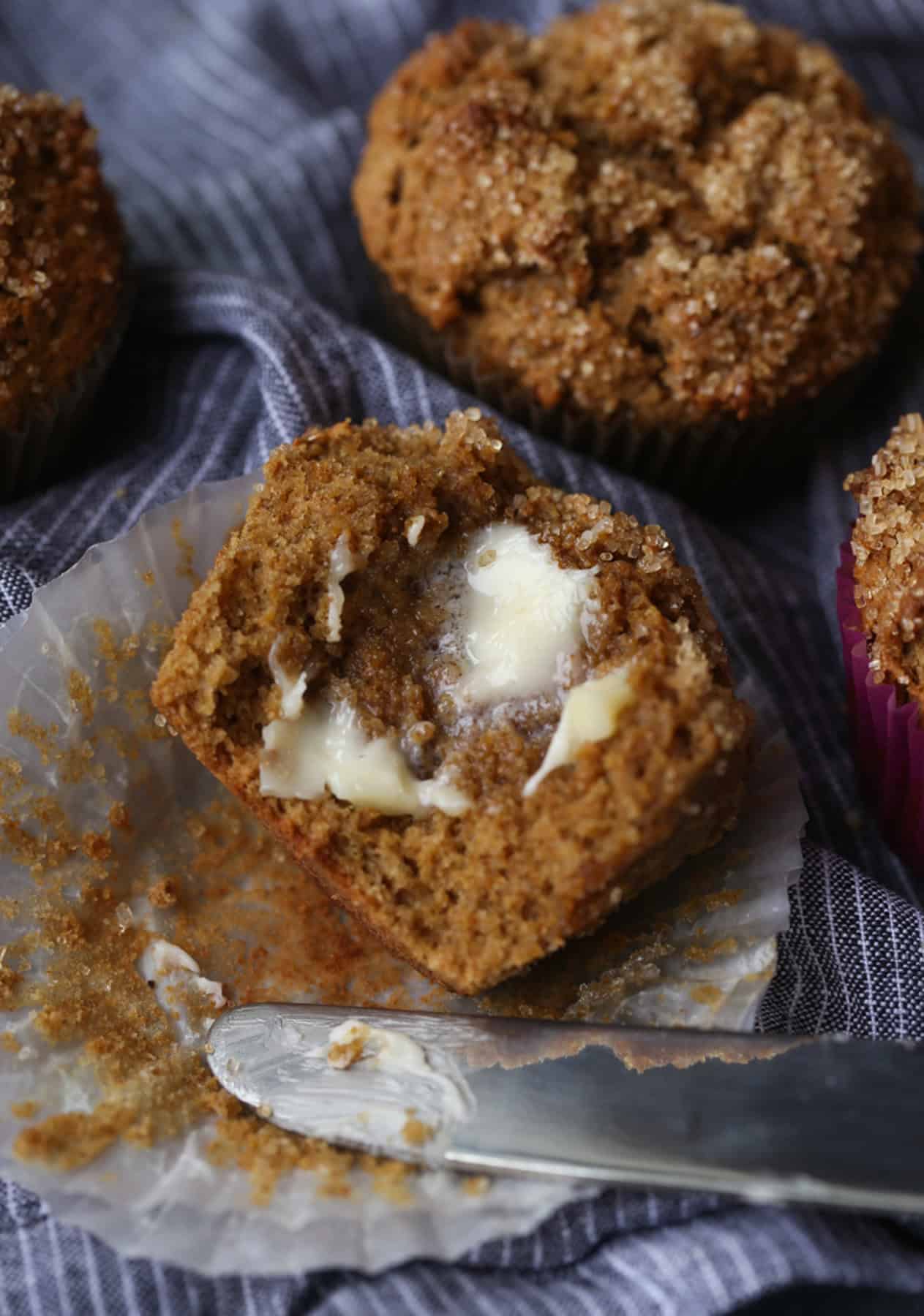 One half of a bran muffin spread with butter next to the tip of a butter knife and a whole muffin.