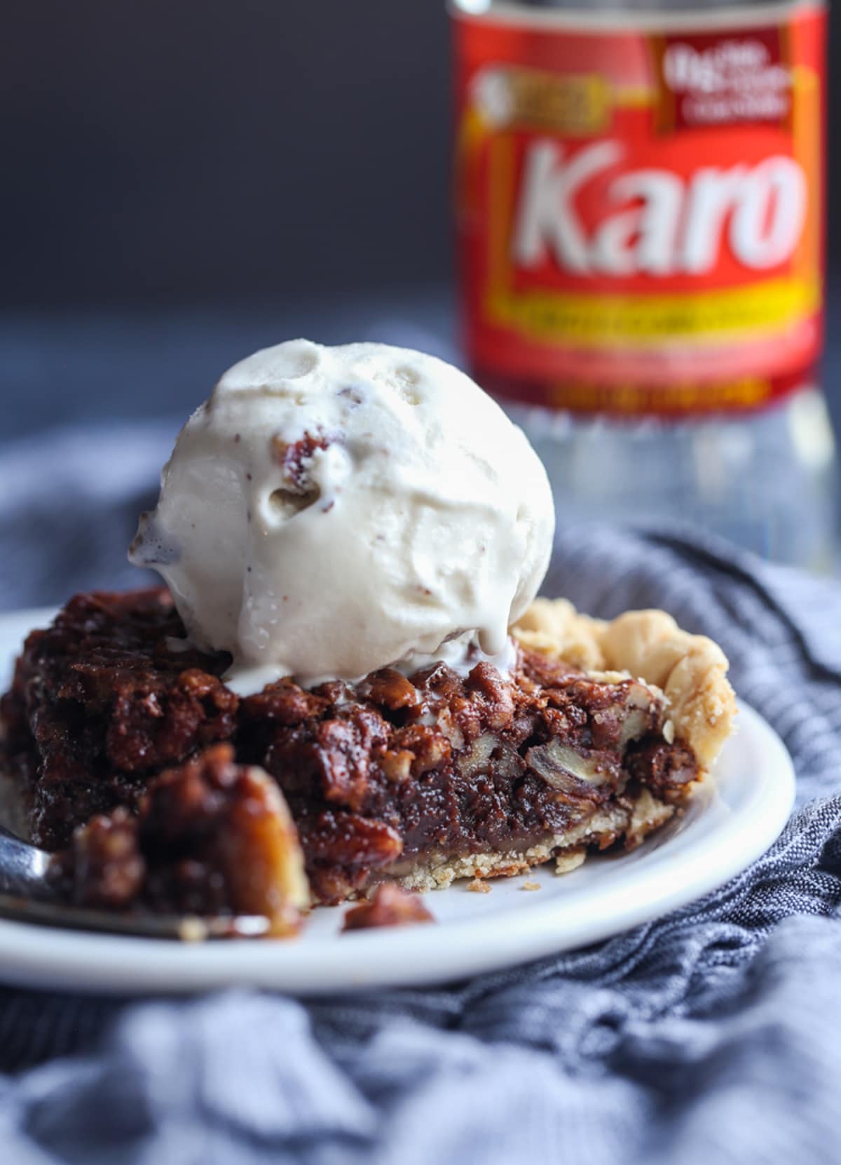 A slice of chocolate pecan pie on a white plate with ice cream and a bottle of Karo syrup in the background