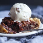 A slice of chocolate pecan pie on a plate topped with vanilla ice cream with a forkful taken out.